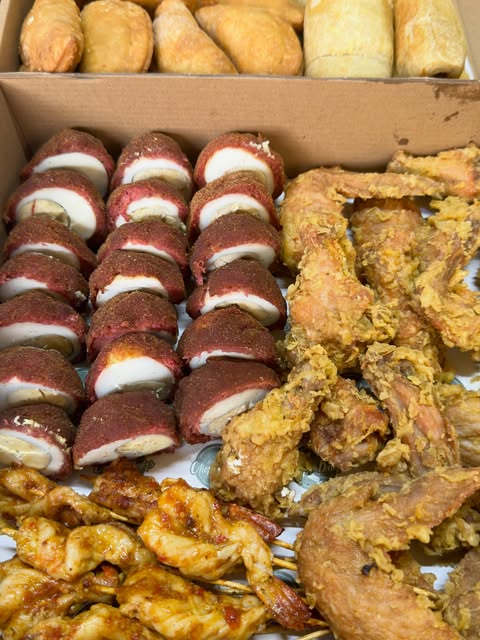 An open event box packed with red-crusted scotch eggs, golden fried chicken wings, samosas and sausage rolls, with skewers of peppered chicken alongside