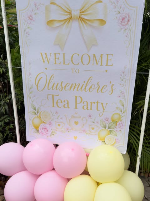 A styled tea-party welcome sign in gold lettering, framed by pink and lemon balloons at an event catered in Abuja